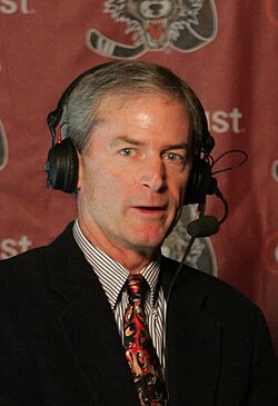 A candid bust photo of a white man with grey hair looking into the camera; he is wearing a shirt, tie, jacket, and large headset