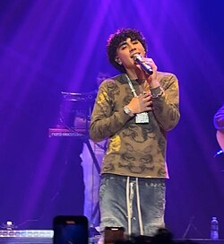 DannyLux performing at Syracuse University on October 19, 2024. He is wearing an army-green shirt, a chain with diamond encrusted castle, blue pants, and diamond earrings