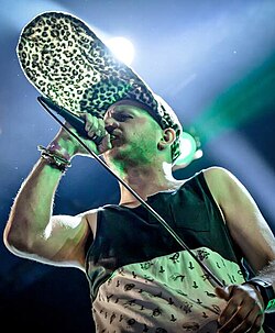 White male standing onstage, holding microphone to his mouth, wearing tank top and unusual hat with brim