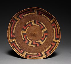 Round Tlingit twined basket tray, late 19th century, woven from spruce root and sea lyme-grass, with concentric geometric bands in brown, black, red, and yellow pigment.