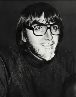 A monochrome photo of a short-bearded man with medium-length hair, wearing glasses with thick black rims.