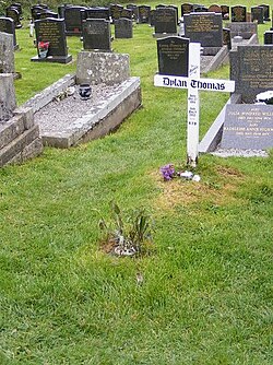 A simple white cross engraved with a memorial message to Thomas stands in a grave