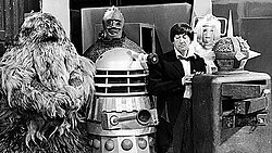 A group shot consisting of: a furry creature; a reptilian figure wearing strange armour; a mechanical, one-eyed robotic creature; a black-haired man wearing a suit and bowtie staring at the robotic creature; a metallic humanoid creature; a boxy creature with a round head, spikes jutting out of all sides of the head