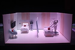 A hand-made model of a hospital room separated by a curtain, with grapes standing in for patients. On the left of the curtain, there are two "patients" wearing surgical masks, one in a bed connected to an electrocardiogram and one sitting down on a chair besides it. On the right, there is a surgical bed where the da Vinci Surgical System performs surgery.