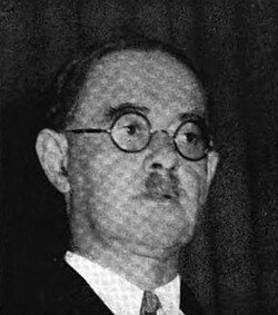 Photograph of a man with circular glasses and a toothbrush mustache