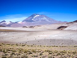 A low cone with a black tongue rises above white, sparsely vegetated terrain