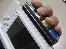 An accessory with four buttons connected to the bottom of an opened gaming handheld. A person's hand runs alongside the handheld's back with fingers placed on the buttons.