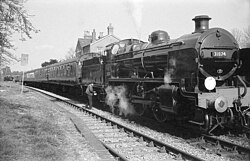 A side-and-front view of a 2-6-0 steam locomotive about to depart a railway station. The locomotive features smoke deflector plates either side of the boiler and there is a crew member oiling the motion. It is hauling four carriages.
