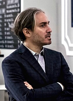 white caucasian male in suit, around 30 years old, appears concerned with arms crossed, dark hair with fine blond highlighted fringe