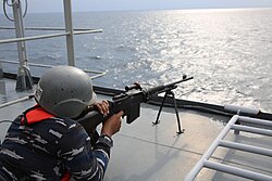 A sailor in naval attire fires a light machine gun from the deck of a vessel, aiming over calm water on a sunny day.