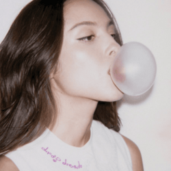 Rodrigo chewing on gum and blowing a bubble; she is wearing a white tank top and the words "drop dead" are embroidered in pink along the neckline of the shirt