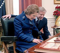 Margaret Thatcher seated behind the Resolute desk with Jimmy Carter looking over her shoulder as she read the plaque installed on the desk.