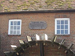 Brick building with tiled roof and two square windows, below which is a water wheel. Between the windows there is a wooden plaque, which reads "Cobham Mill Restored 1993 by Cobham Mill Preservation Trust and the National Rivers Authority".