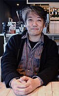 Portrait of a Japanese man sitting at a table wearing a black jacket and grey flannel shirt.