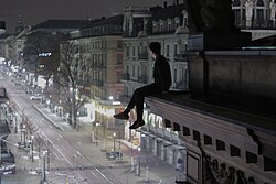 A person sits on the ledge of a building looking down at the street below