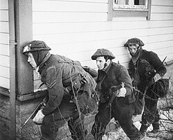 A black and white photograph of three British soldiers advancing beside a wooden house. The lead soldier is carrying a Thompson submachine gun and is peering around the corner of the house, while the other two are carrying rifles.