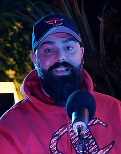 A man with a long beard, black cap, and red hoodie in front of a microphone