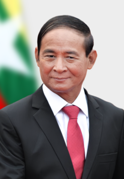 Photograph of Win Myint being dressed in a black suit jacket, white shirt, and red tie. The Myanamr flag is also seen in the background.