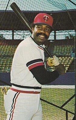 A black man in a white baseball uniform with a red cap stands in a left-handed batting stance.