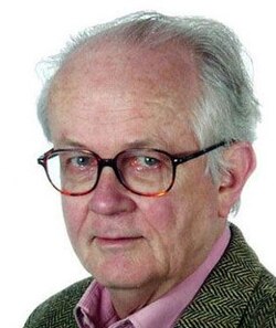 Colour three-quarter profile publicity shot, of clean-shaven, bespectacled half-smiling man, with receding short white hair, brushed back