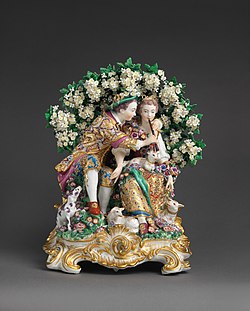 A porcelain figurine of a young couple sitting under a flowering tree, with the boy teaching the girl to play a flute, surrounded by sheep and dogs.