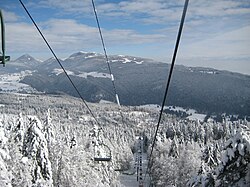 A winter mountain landscape in France with a chairlift ascending over snow-covered forests and distant peaks under a blue sky.
