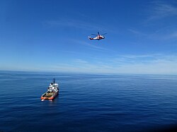 Red helicopter flying above a supply vessel during offshore support operations on calm sea.