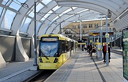 Two yellow trams sit besides one another on parallel tracks, with the one on the left slightly closer to the camera which is on the ground in the road. Both have their pantographs up which are touching contact wires. On either side there are people walking on the pavement.