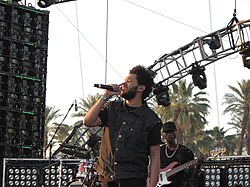 The Weeknd wearing a button-up, sleeveless shirt covering a normal black t-shirt. He is performing on a stage, singing into a microphone, with palm trees in the background.