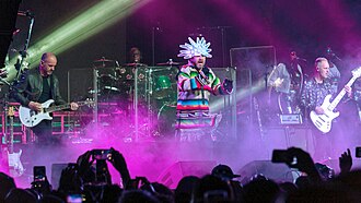 The band Jamiroquai performing on stage at Coachella 2018. At center is vocalist Jay Kay wearing a colourful Native-American shirt with frills around the waist and a white LED head-dress. Also present are a guitarist, a bassist, a drummer and two female backing vocalists. The band is engulfed in roughly waist-level stage fog coloured purple by stage lighting.