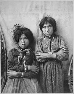 Black-and-white photograph of two Tlingit girls standing side by side near the Copper River, Alaska, in 1903, wearing patterned dresses, jewelry, and nose rings, with their arms crossed.