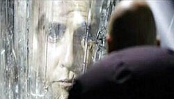 The face of a man appears in a crystalline wall, while a bald man with his back to the camera looks at him.