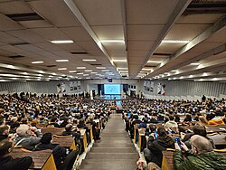 Wide angle photograph of the nearly full Jansen auditorium at ULB from the upper stairs looking down. Depicting people queueing on the sides to find an empty seats while the speaker prepares to give the FOSDEM 2025 Closing Talk.