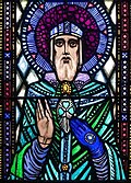 St. Patrick, with a halo, and wearing a bishop’s mitre, places his hand upon a shamrock below the clasp of his cloak.