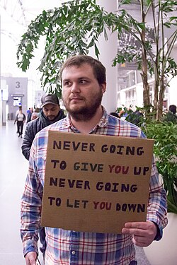 A person holds a cardboard sign with the words, "Never going to give you up, Never going to let you down".