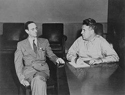 Groves sits at a completely clean desk. Chadwick, seated next to him, looks on.