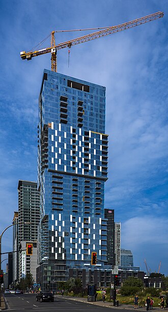A tall bluish glass-sheathed rectilinear tower with a tower crane atop, rising from an area behind a vacant lot, in front of which is an intersection with red traffic lights. A blue sky speckled with small clouds is in the background.