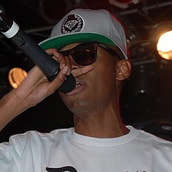 A black male with sunglasses, a white shirt with text “Death,” and a green and white snapback speaking into a microphone