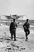 Two armed men, one with a rifle with a bayonet attached and the other carrying a submachine gun, walking along the water's edge below a line of cliffs.