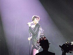 Photograph of a woman who sings on stage. She wears a dark leather blouse and a short skirt, and light tights.