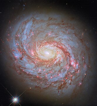 "A close-up of a spiral galaxy, seen face-on. Its center is a bright glowing From the sides of the galaxy's core emerge spiral arms which wind through the round disc of the galaxy, filled with shining pink spots where stars are forming and more dark-red dust. Some faint stars can be seen around the galaxy, as well as a particularly bright star in the lower left of the image."