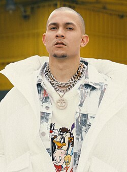Puerto Rican producer Tainy posing for a photo, sporting a white jacket, a buttoned shirt, a Chanel necklace, and a Looney Tunes shirt.