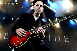 slightly blurry image of Billy Howerdel wearing dark clothing, playing a red electric guitar onstage, "Ashes Divide" written in large letters on the backdrop