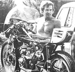Black and white image of a man stripped to the waist in strong sunshine, alongside a part dismantled racing motorcycle