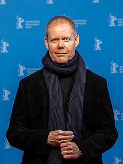 A middle-aged man with reddish hair wearing a black jacket and a dark blue scarf, photographed against a blue background
