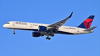A mostly white Boeing 757 with blue and yellow trim preparing for landing against a grey sky.
