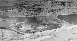 Black and white photograph of a ship in a bay. Smoke is drifting up from the land.