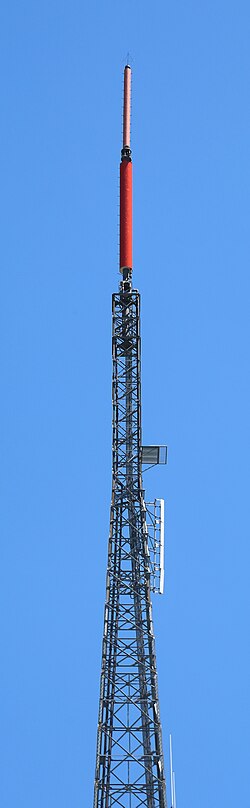 A gray lattice tower, set against a blue sky. A small pink cylindrical antenna and a larger red cylindrical antenna top the structure.