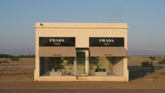 A small one story, one room adobe building with two large plate glass windows. Purses and shoes are sparsely and methodically placed inside.