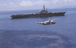 A color photograph of an helicopter flying above an aircraft carrier
